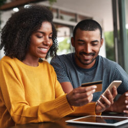 Young african man looking at his girlfriend using credit card and smartphone for shopping online in cafe
